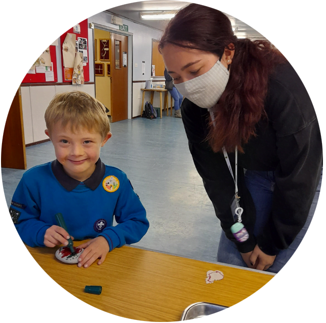 A young boy smiling while doing a craft while a volunteer wearing a mask supervises him
