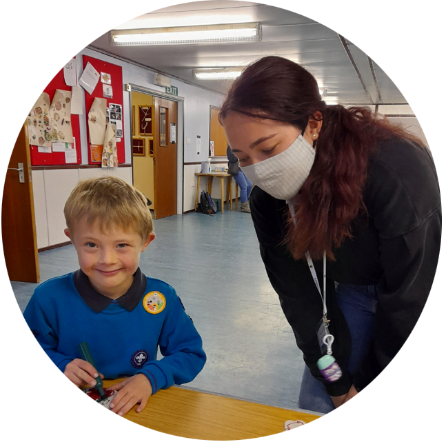 A young boy smiling while doing a craft while a volunteer wearing a mask supervises him
